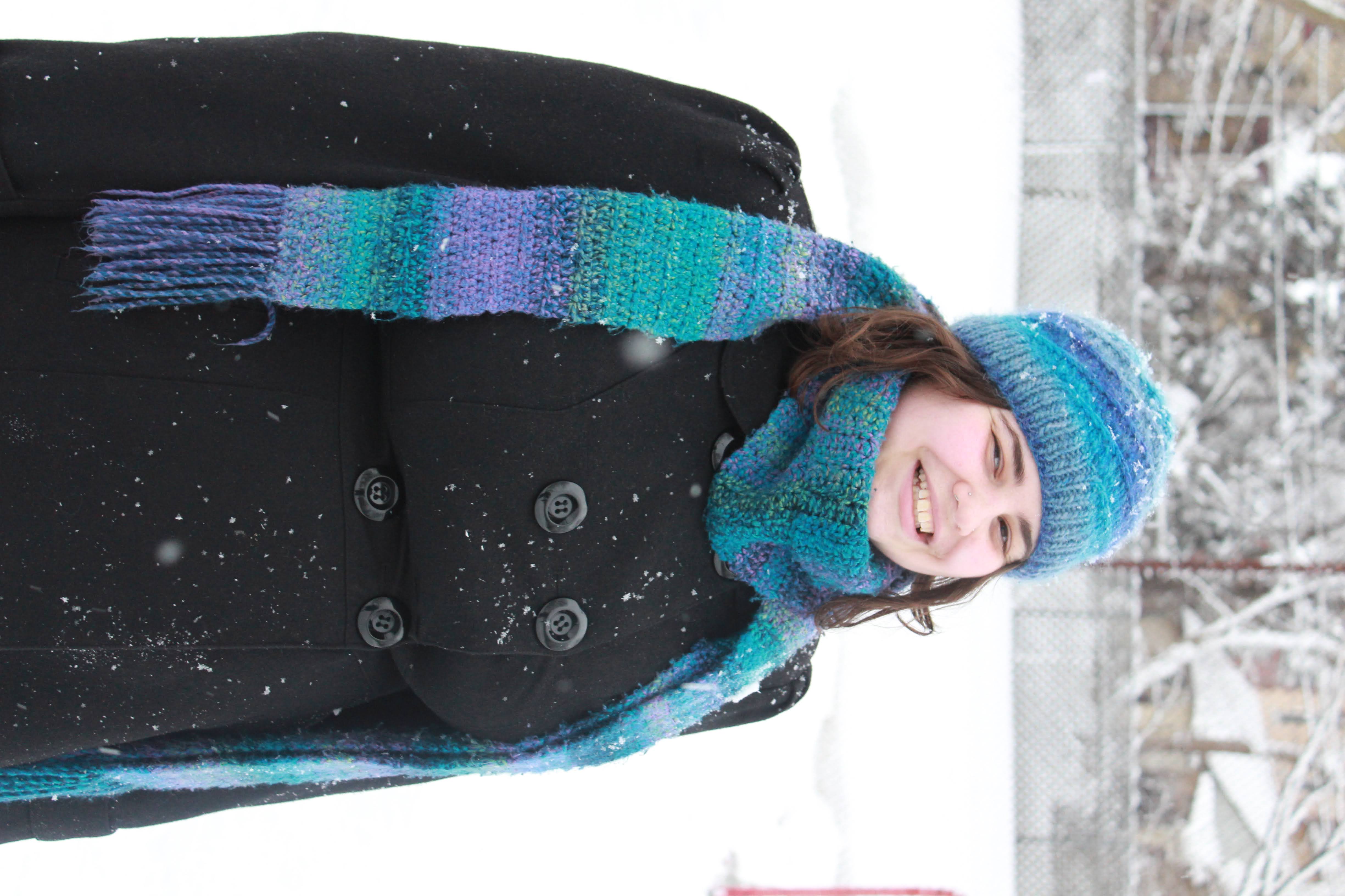 A photograph of Cassie smiling. It’s winter and they are wearing a black coat, and blue and purple hat and scarf they knitted.